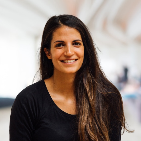 Young woman with long brown hair, wearing a black top, smiling at the camera in an indoor setting.